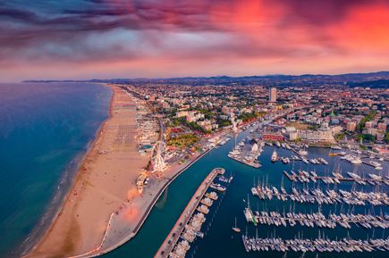 Blick über Rimini im Morgenrot. Auf der linken Seite das Meer, davor der Sandstrand. Rechts davon ein kleiner Hafen und im Hintergrund die Stadt.