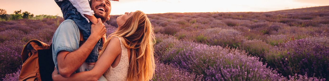 Ein verliebtes Paar in Weiß schlendert Hand in Hand durch ein blühendes Lavendelfeld, umgeben von violetten Blüten, unter einem klaren, sonnigen Himmel.