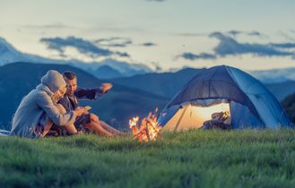 Zwei Freunde sitzen vor einem Zelt am Lagerfeuer in den Bergen bei Sonnenuntergang.