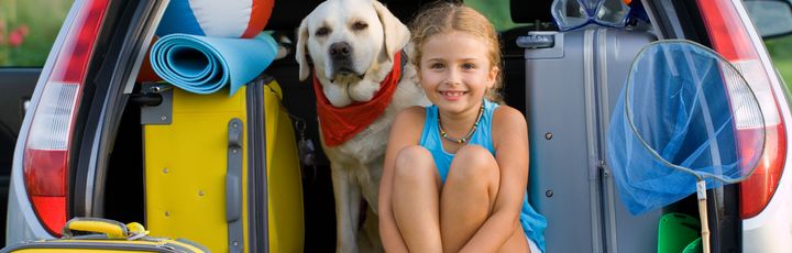 Zwei Mädchen und ein Hund sitzen fröhlich im geöffneten Kofferraum eines Autos.