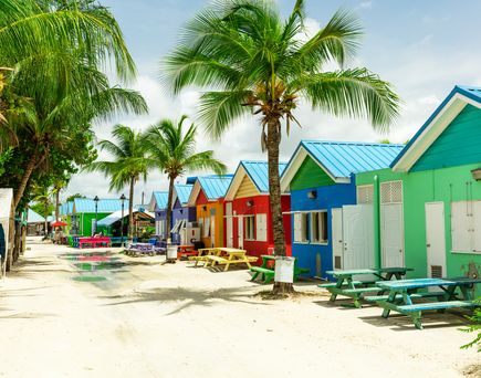 Barbados. Palmen, Sandstrand und daneben viele kleine bunte Häuser, direkt im Sand gebaut. Blauer Himmel und blaues Meer.