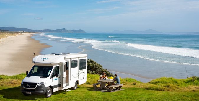 Camper parkt an Te Ārai Regional Park auf einer Wiese vor dem Strand in Mangawhai in Neuseeland. Vor dem Camper sitzen zwei Personen an einem Tisch und unterhalten sich.