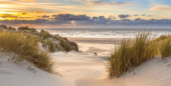 Ein typisches Naturbild Hollands. Dünen, Sand, etwas Gras und in der Ferne das Meer am blauen Himmel mit Sonnenuntergang. Menschenleer.