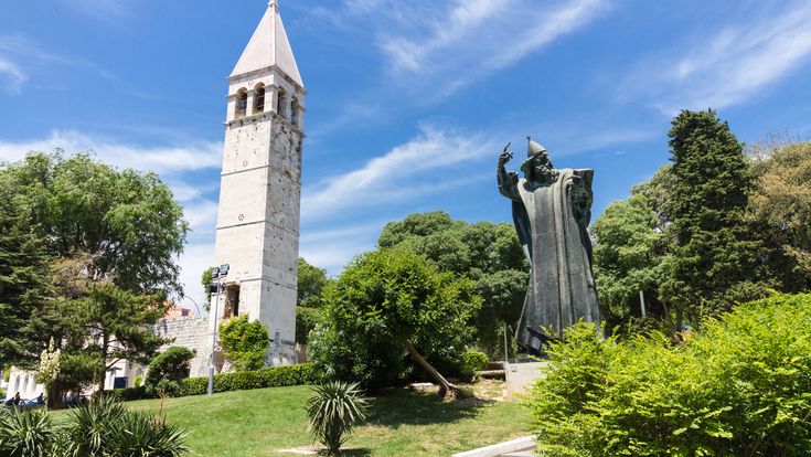 Eiserne Statue von Gregor von Nin, dahinter eine Kirche.  Umgeben von Wiese und Bäumen, blauer Himmel.
