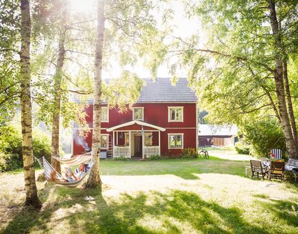 Ein kleines, rotes Holzhaus mit einem weißen Zaun und einem grünen Garten, umgeben von Bäumen. Die Szene wirkt gemütlich und einladend.