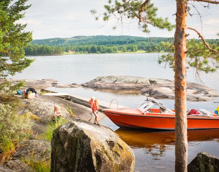 Eine rote Fischerhütte oder ein Bootshaus am Wasser, umgeben von Bäumen. Die Szene ist ruhig und sonnig.
