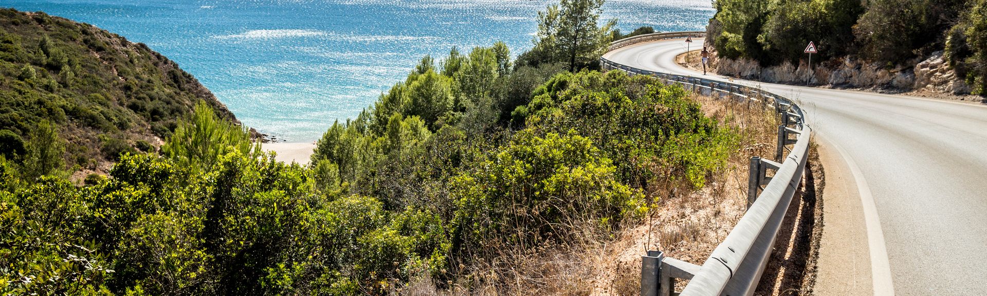Eine malerische Küstenstraße, die Estrada da Arrábida, schlängelt sich entlang der Küste der Algarve in Portugal, umgeben von grüner Vegetation und dem blauen Atlantik unter einem klaren blauen Himmel.