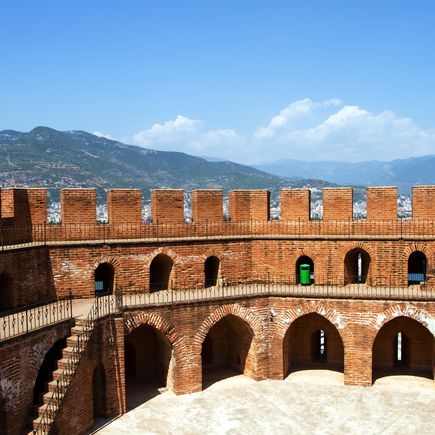 Die Rote Turm (Kızıl Kule) in Alanya, Türkei, eine beeindruckende achteckige Festung aus rotem Backstein, die sich majestätisch über der Küste erhebt, mit dem Meer im Hintergrund und einem bewölkten Himmel.
