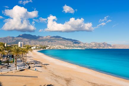 Strand Playa del Albir an der Costa Blanca in Spanien, mit türkisblauem Meer, einem sandigen Strandabschnitt, Palmen am Promenadenrand und Bergen im Hintergrund