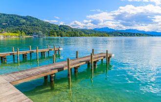 Zwei Holzstege ragen in das türkisblaue Wasser des Wörthersees in Österreich, umgeben von grünen Hügeln.