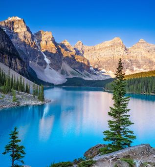 Kanadische Landschaft mit hellblauem See und Berge im Hintergrund, die von der Sonne angestrahlt werden