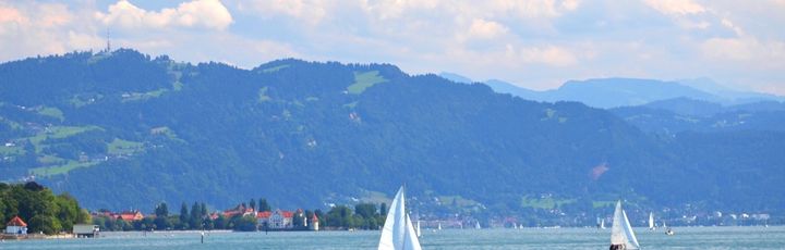 Ein Panoramablick auf den Bodensee, umgeben von malerischen Städten und grünen Hügeln, mit Booten auf dem Wasser und einem klaren blauen Himmel.
