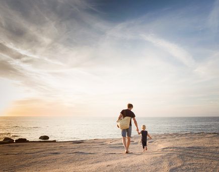 Eine einzelne Person geht am Strand entlang, während die untergehende Sonne den Himmel in warme Orangetöne taucht und sich im ruhigen Meer spiegelt.