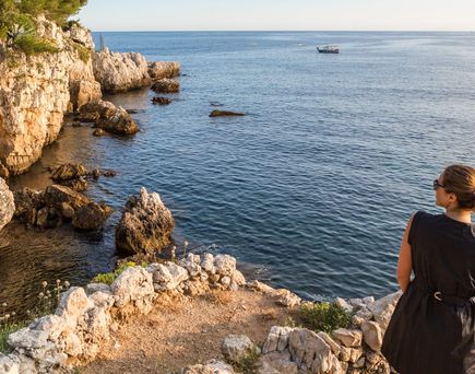 Eine Person sitzt auf einer felsigen Klippe und blickt auf das weite, ruhige Meer. Die Landschaft ist sonnig und friedlich.