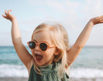 Kleines Kind mit Sonnenbrille steht am Meer und streckt die Arme nach oben