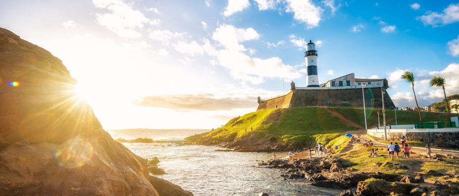  Der historische Farol da Barra Leuchtturm in Salvador, Brasilien, steht auf einer Klippe über einem felsigen Strand, umgeben von Bäumen und dem Ozean.