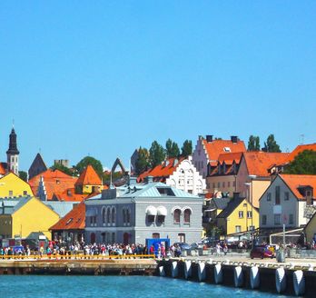 Blick auf die bunten Häuser von Visby, ein Steg am Wasser, Cafe und Sitzmöglichkeiten dort. Blauer Himmel.
