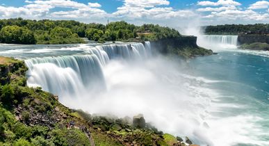 Zu sehen sind die beeindruckenden und weitläufigen Niagara Fälle, inmitten von hohen Bäumen. Blauer Himmel mit einigen kleinen weißen Wolken am Himmel.