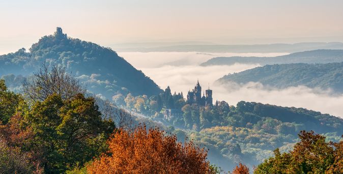 Das majestätische Schloss Drachenburg thront auf einem bewaldeten Hügel im Siebengebirge, mit dem Drachenfels im Hintergrund, während Nebel über den unteren Lagen der Landschaft schwebt.
