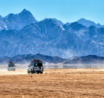 Mehrere weiße Geländewagen fahren im Konvoi durch die staubige Wüste vor einer eindrucksvollen, bläulichen Bergkette.