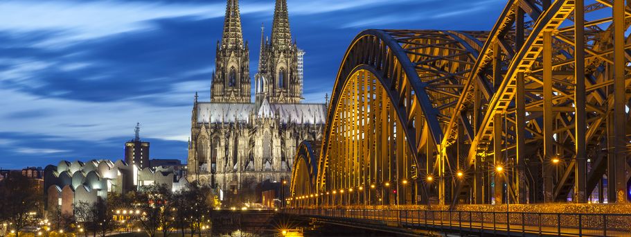 Der Kölner Dom und die Hohenzollernbrücke bei Nacht, beleuchtet, spiegelt sich im Rhein.