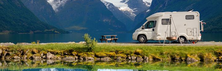 Weißer Campervan steht am Ufer eines Sees in Norwegen, spiegelt sich im Wasser, umgeben von grünen Bergen.
