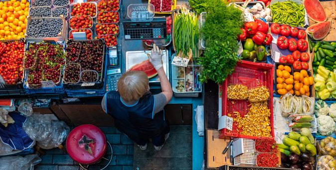 Ein reich gedeckter Obststand, Blick von oben. Zu sehen sind Obst und Früchte. Die Verkäuferin wiegt grade mit Handschuhen ein Stück Wassermelone auf einer Metallwage ab.