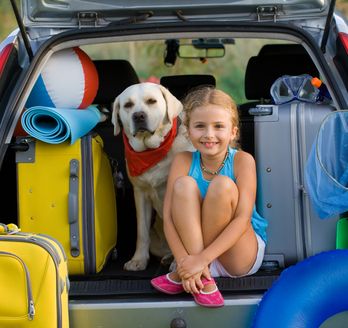 Zwei Mädchen und ein Hund sitzen fröhlich im geöffneten Kofferraum eines Autos.