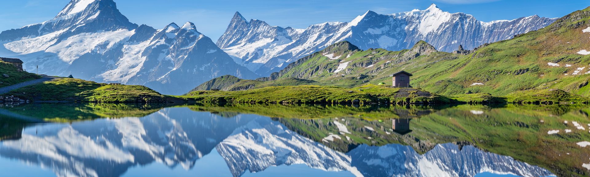Der Bachalpsee in den Schweizer Alpen, der die schneebedeckten Gipfel des Wetterhorns, Schreckhorns und Finsteraarhorns sowie des Grindelwaldgletschers in seiner spiegelnden Oberfläche reflektiert. Am Ufer steht eine kleine Holzhütte auf einer grünen Wiese unter einem klaren blauen Himmel.
