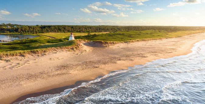 Zu sehen ist das wilde Meer, links daneben etwa Strand und dann die grünen Dünen. Einsam steht ein Leuchtturm.
