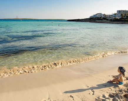 Ein Sandstrand mit Felsen und Meer, ein Baby sitzt im Sand mit blauem Kopftuch.