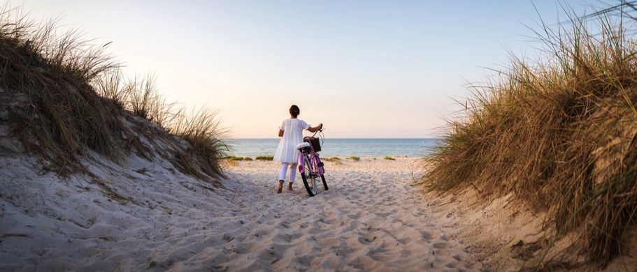 Mutter mit Kind und Fahrrad geht durch eine Sanddüne an der Ostsee Richtung Meer im Abendlicht.