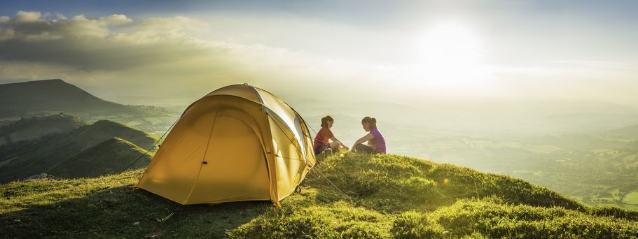 Zwei Personen sitzen neben einem gelben Zelt auf einem grasbewachsenen Hügel bei Sonnenuntergang mit weiter Aussicht ins Tal.