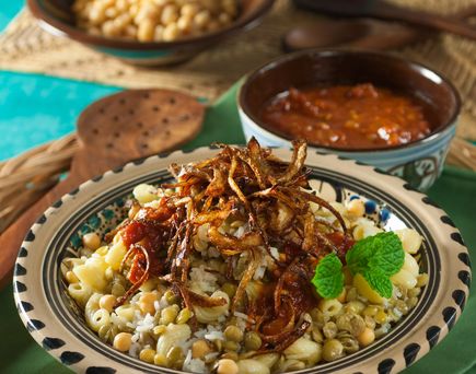 Ein Teller Koshari, ein traditionelles ägyptisches Gericht mit Reis, Makkaroni, Linsen, Kichererbsen und knusprigen Röstzwiebeln, daneben Schalen mit Soßen.