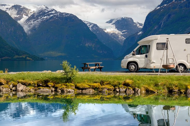 Weißer Campervan steht am Ufer eines Sees in Norwegen, spiegelt sich im Wasser, umgeben von grünen Bergen.