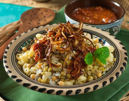 Ein Teller Koshari, ein traditionelles ägyptisches Gericht mit Reis, Makkaroni, Linsen, Kichererbsen und knusprigen Röstzwiebeln, daneben Schalen mit Soßen.