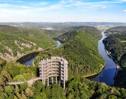 Eine spektakuläre Luftaufnahme der Saarschleife von der Cloef aus, die den mäandernden Fluss Saar zeigt, der sich durch eine bewaldete Hügellandschaft schlängelt, unter einem blauen Himmel mit einigen Wolken.