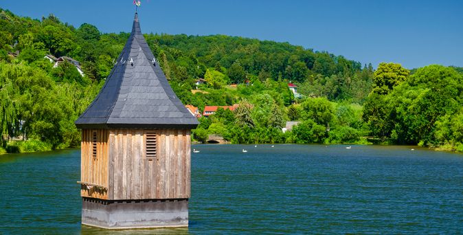 Ein beeindruckender Nachbau des ehemaligen Kirchturms Waldeck ragt aus den Wassern des Edersees empor, umgeben von einem weitläufigen See und bewaldeten Ufern unter einem klaren Himmel.
