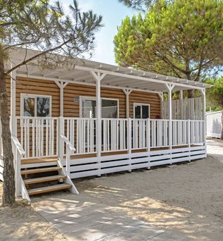 Modernes Mobilheim mit Holzfassade und überdachter Veranda in einem schattigen Kiefernwald auf sandigem Boden.
