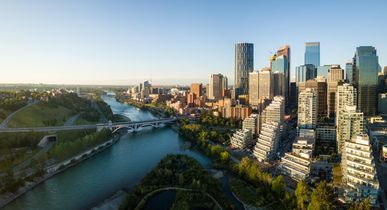 Die Stadt Calgary. Viele Hochhäuser, teilweise direkt am Fluss mit kurzer Brücke, blauer Himmel, Sonnenuntergang. In der Ferne im Nebel kann man die Berge, die Rocky Mountains, erkennen.
