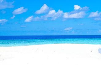 Ein unberührter Sandstrand mit einem einzelnen Sonnenschirm und Liegestühlen unter einem strahlend blauen Himmel. Das türkisfarbene Meer ist ruhig.