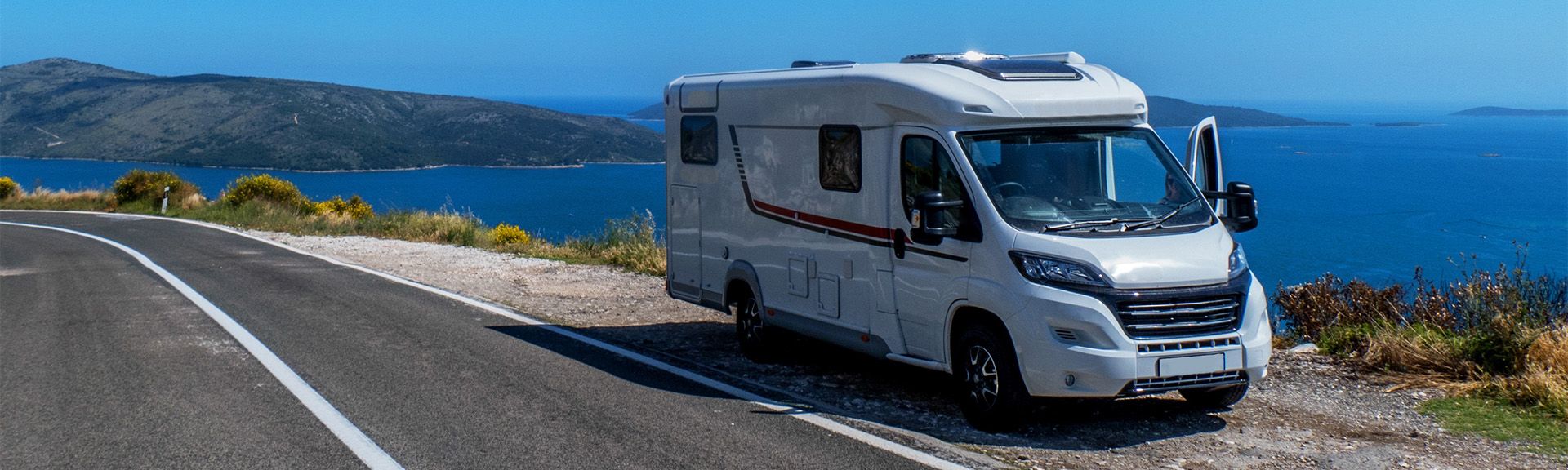 Ein Reisemobil fährt auf einer kurvenreichen Küstenstraße am Meer entlang, umgeben von grünen Klippen und einem blauen Himmel mit Schäfchenwolken.