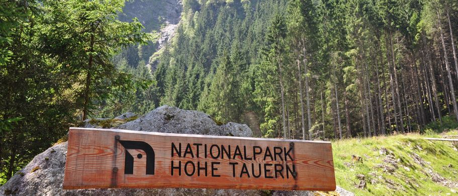 Nationalpark Hohe Tauern. Vorne steht ein entsprechendes Holzschild mit Aufschrift, dahinter der Wald und die Berge.
