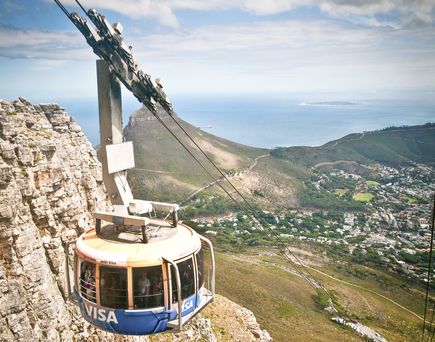 Eine Seilbahnkabine fährt den Tafelberg in Kapstadt, Südafrika, hinauf oder hinunter, mit einer weiten Aussicht auf die Stadt und das Meer unter einem klaren Himmel.