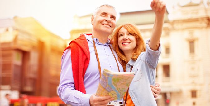 Lächelndes Paar mittleren Alters beim Sightseeing in einer sonnigen Stadt; der Mann hält eine Karte, die Frau zeigt begeistert nach oben.
