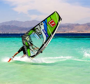Ein Windsurfer auf dem türkisfarbenen Wasser, der ein grünes Segel mit einer roten und weißen Grafik auf einem sonnigen Tag benutzt.
