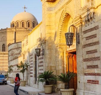 Die historische Hängende Kirche (Church of St. George) in Kairo, mit ihrer markanten Architektur und einem runden Kuppelbau im Hintergrund, bei hellem Tageslicht.