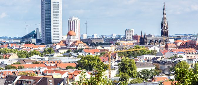 Skyline von Leipzig mit historischen und modernen Gebäuden, darunter das City-Hochhaus, Kirchen und grüne Baumkronen im Vordergrund.