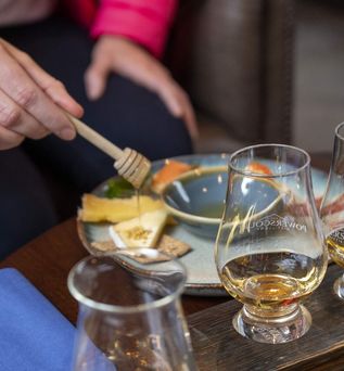 ine Nahaufnahme von drei Glencairn-Whiskygläsern, gefüllt mit goldener Flüssigkeit, die auf einer hellbraunen Holzoberfläche stehen. Im Hintergrund ist eine Hand zu sehen, die Honig auf einen kleinen Teller mit Käse und Gebäck träufelt.