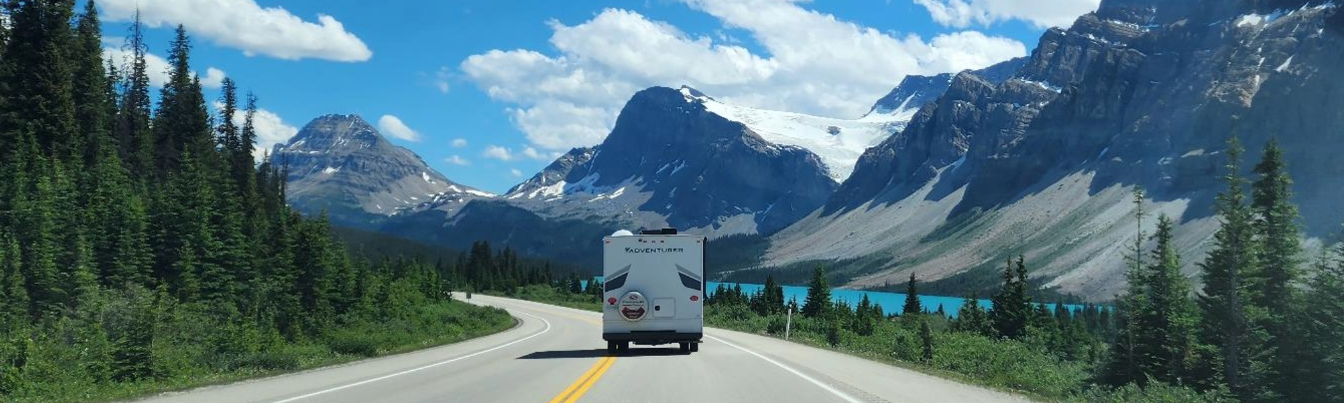 Eine atemberaubende Panoramaansicht des Icefields Parkway im Banff Nationalpark, Kanada, mit einer Straße, die sich durch eine majestätische Berglandschaft schlängelt, umgeben von schneebedeckten Gipfeln und einem weiten blauen Himmel.